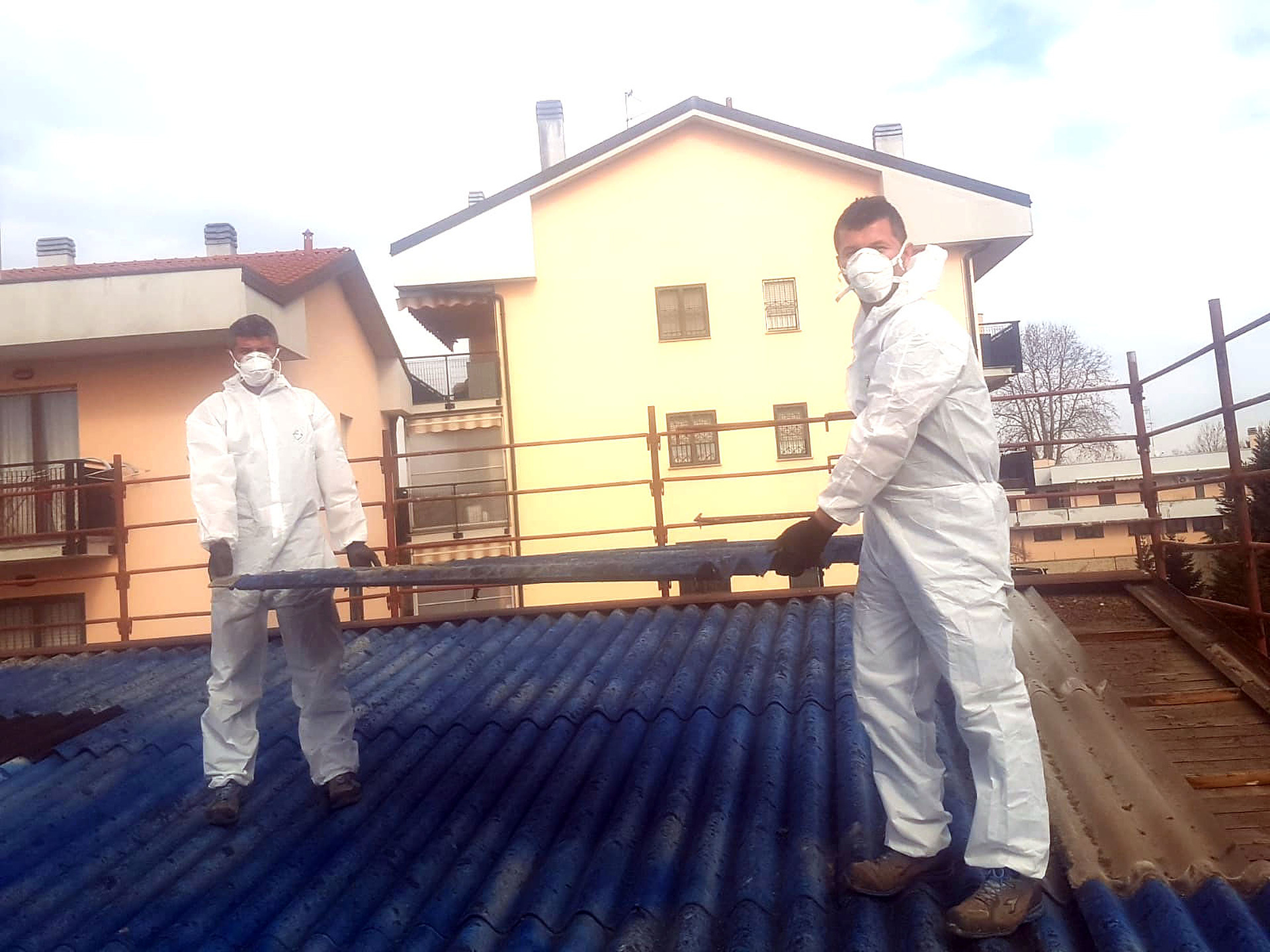 Due operai di F.lli Lulja in tuta protettiva bianca e mascherina sollevano una lastra ondulata blu durante la rimozione di una copertura in cemento amianto