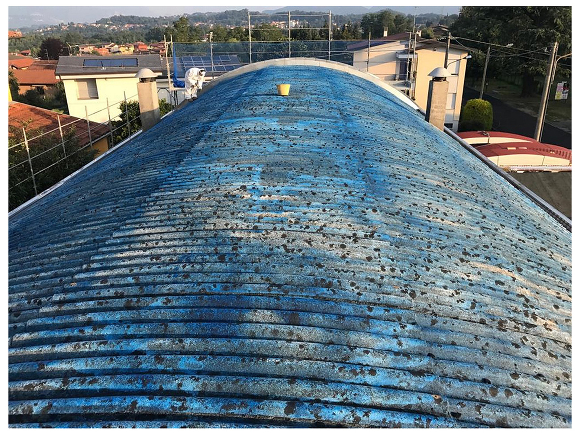 Copertura curva in lastre ondulate di cemento amianto di colore blu, con un operaio in tuta protettiva in fondo al tetto