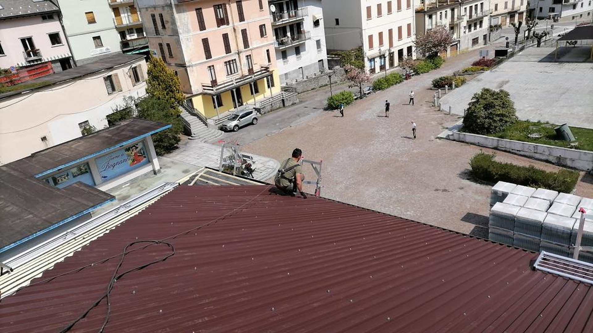 Operaio al lavoro su una copertura in lamiera grecata rosso scuro, affacciata su una piazza di paese